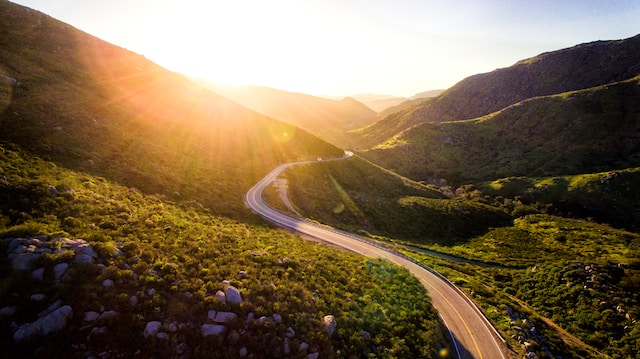 A road leading through green mountains, with the sun peaking over the horizon.